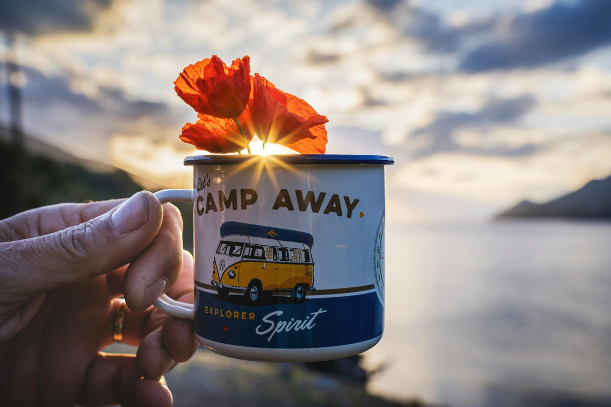A man holds a camping mug in his left hand, with a picturesque view of towering cliffs meeting the ocean in the background.