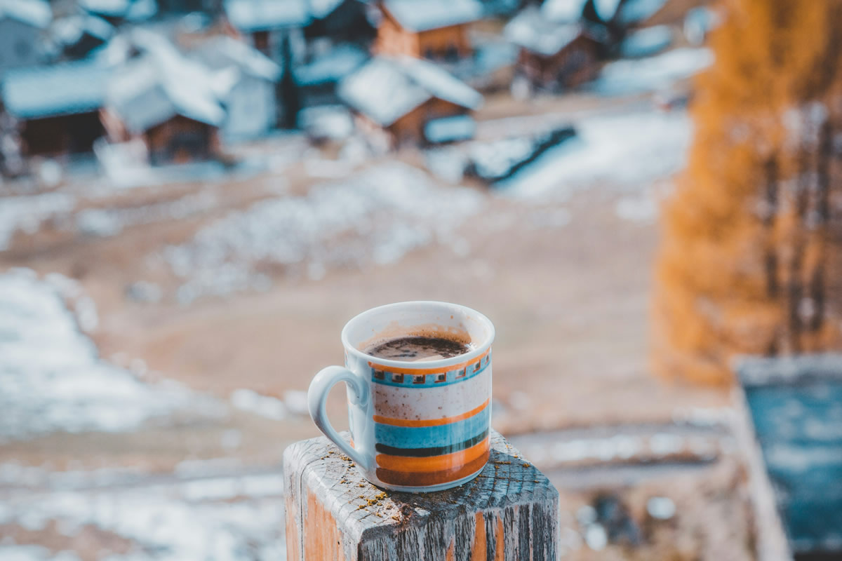 A mug, adorned with picturesque colors, rests on a wooden column. In the background, a stunning winter-fall landscape unfolds.